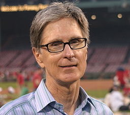 fenway sports group owner john henry wearing a plaid blue shirt and glasses standing in front of a sports pitch