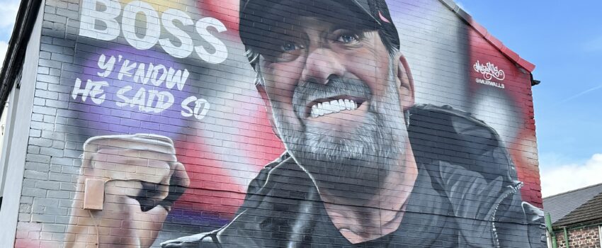 mural of jurgen clop on the side of a house near anfield. klopp is wearing a baseball cap and punching the air with the words 'boss, you know he said so' written to the side.