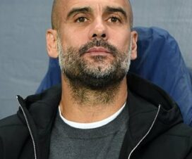pep guardiola wears a white t shirt under a grey jumper with a black coat. he is sitting in front of a champions league hoarding.